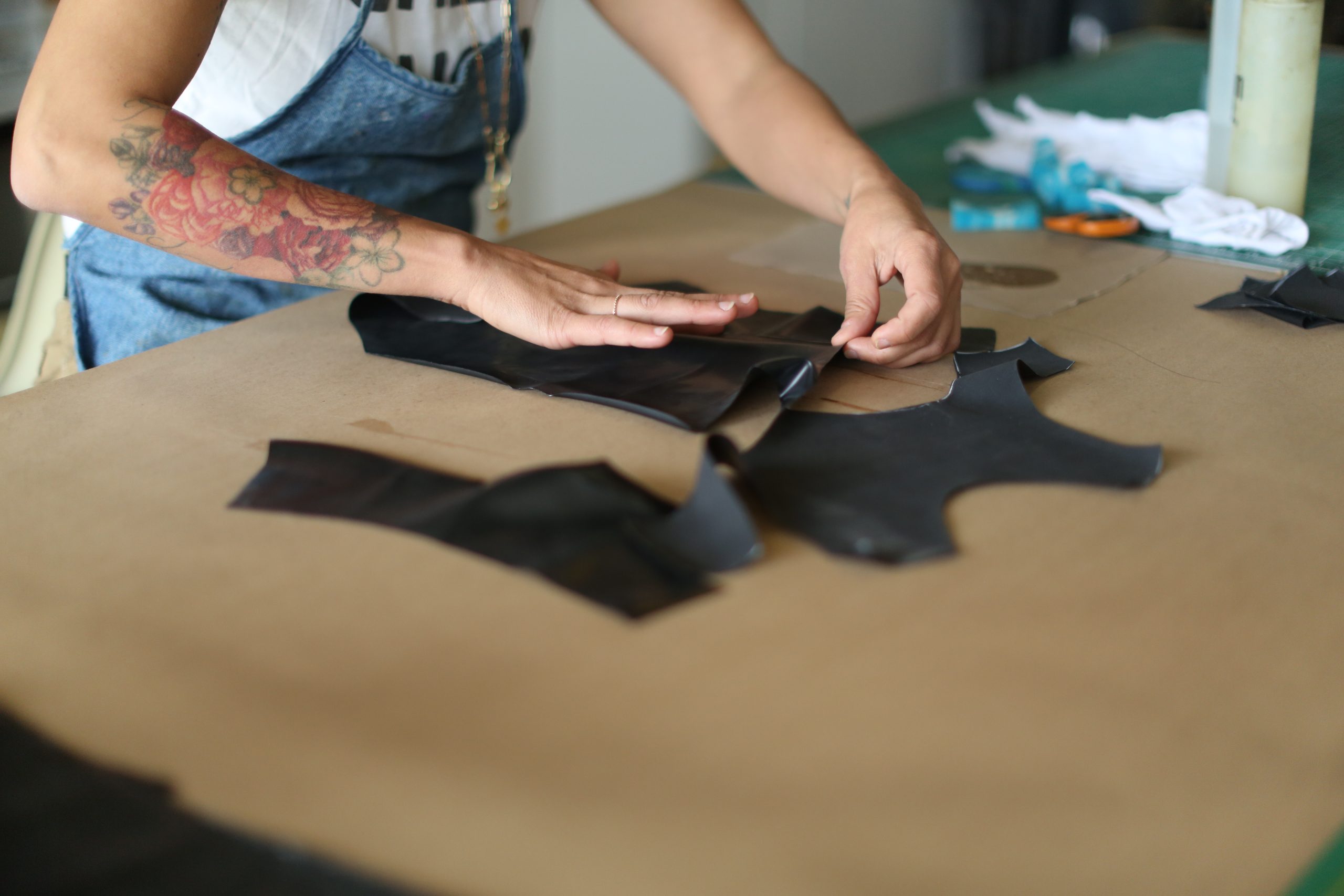 Latex Fashion by Vex A person with a colorful arm tattoo arranges black leather pieces on a brown work surface, preparing materials for crafting. Scissors and other tools are visible in the background.