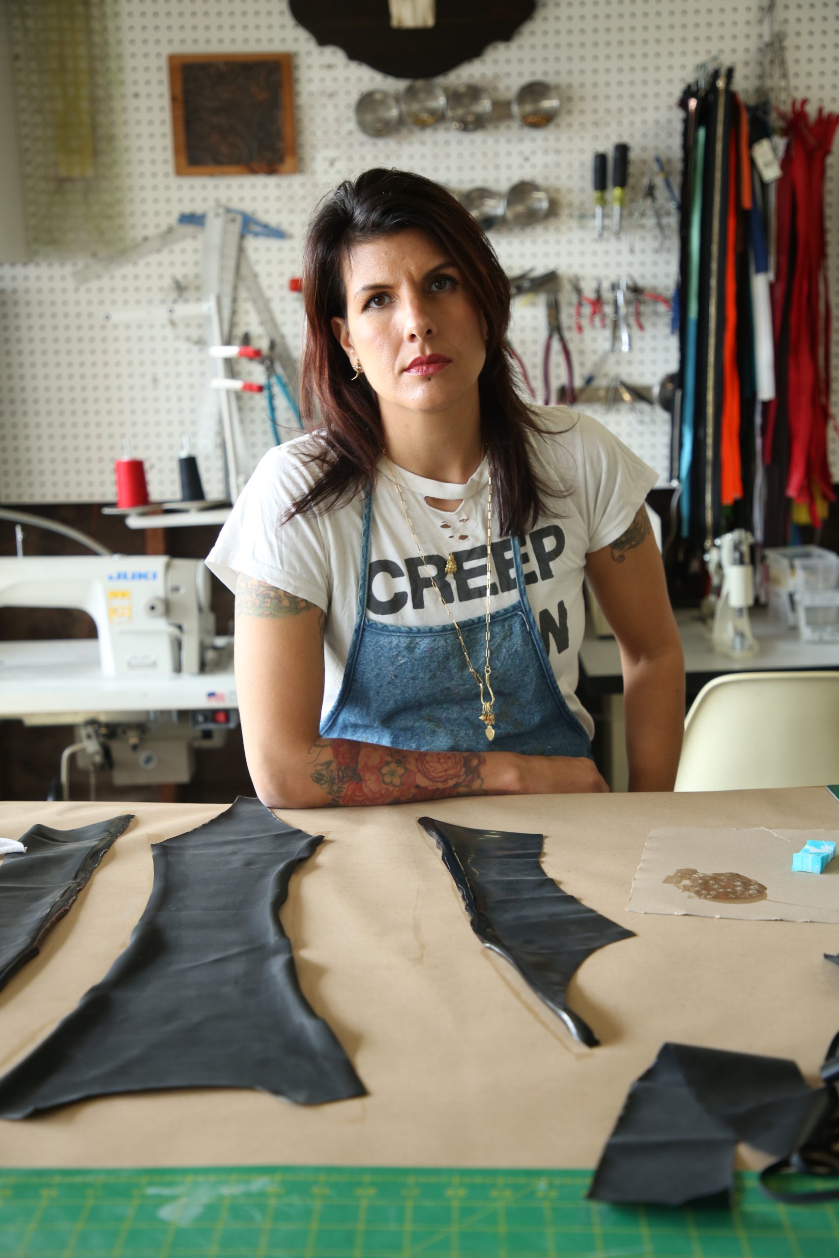 Latex Fashion by Vex A woman with long dark hair wearing a white CREEP ON t-shirt and apron sits at a worktable with leather pieces, sewing tools, and supplies in a workshop setting.